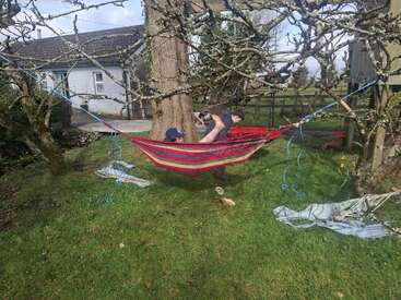 Deux enfants jouent dans un hamac coloré suspendu entre deux arbres dans un jardin verdoyant. À proximité, des couvertures et des chaussures sont éparpillées, et on aperçoit une maison.