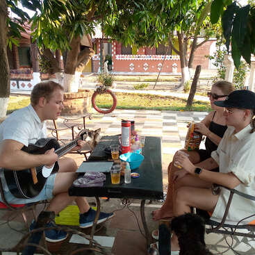 Três pessoas relaxam ao ar livre sob as árvores; uma toca violão enquanto duas desfrutam de lanches e bebidas em uma mesa. A luz do sol passa pelas folhas, criando um ambiente tranquilo.