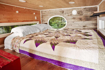Esse loft aconchegante tem uma cama grande com uma colcha colorida, paredes de madeira, janelas ovais e retangulares, travesseiros brancos e luz natural suave entrando.