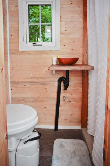 Esse é um banheiro compacto com paredes com painéis de madeira, um vaso sanitário de compostagem, uma pequena janela, balcão da pia de madeira, pia com vaso e cortina de chuveiro branca. Tapete no chão.