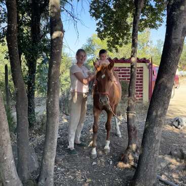 The image depicts a woman and a child standing beside a brown horse with white hooves, in a wooded area with trees and a red brick structure in the background.