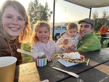 Uma família feliz está sentada ao ar livre em uma mesa de piquenique, saboreando sobremesas doces. A menina mostra a língua, enquanto todos sorriem calorosamente sob a luz do sol.