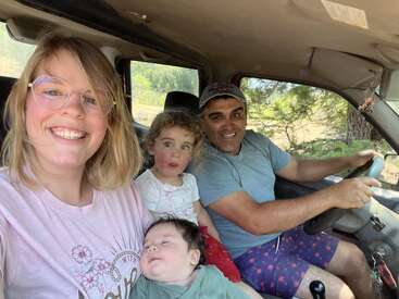 Uma família sorridente de quatro pessoas está sentada dentro de um carro. Dois adultos estão na frente, com seus dois filhos pequenos, aproveitando juntos um dia ensolarado ao ar livre.