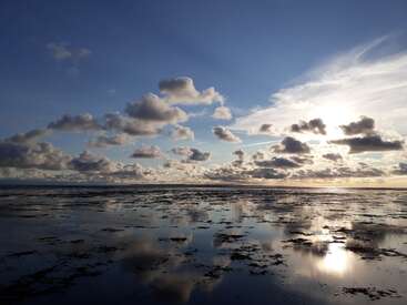 L'image représente une scène de plage sereine avec une vaste étendue d'eau, un ciel bleu avec des nuages et un soleil bas brillant à l'horizon.