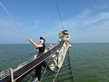 L'image représente une personne debout sur un bateau, saisissant une corde à deux mains, sur un fond serein d'une vaste étendue d'eau et d'un ciel d'un bleu limpide.