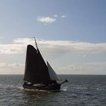 L'image représente un petit voilier naviguant sur l'océan, ses voiles sombres se gonflant dans le vent tandis qu'il glisse sur l'eau agitée.
