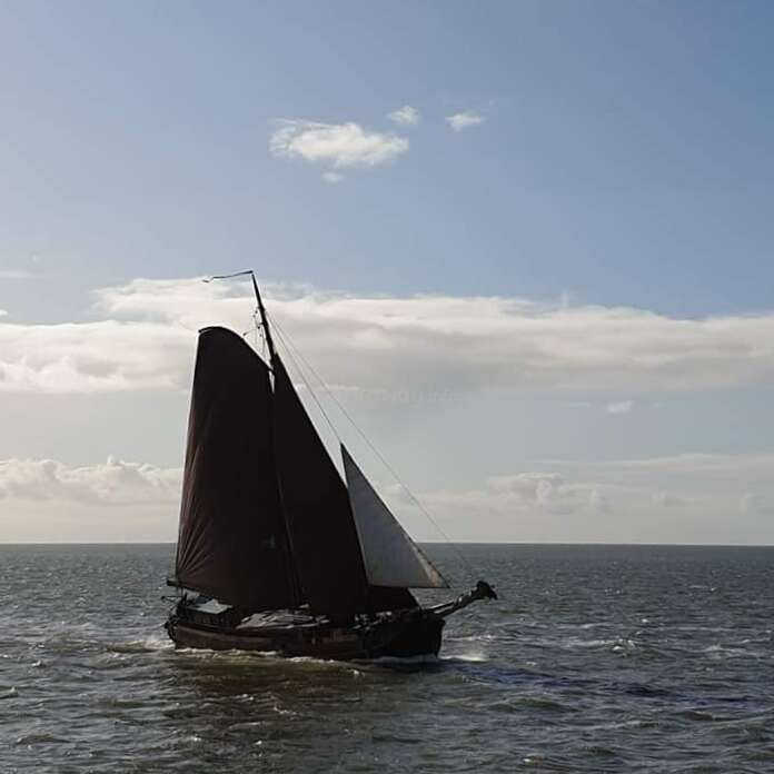 L'image représente un petit voilier naviguant sur l'océan, ses voiles sombres se gonflant dans le vent tandis qu'il glisse sur l'eau agitée.