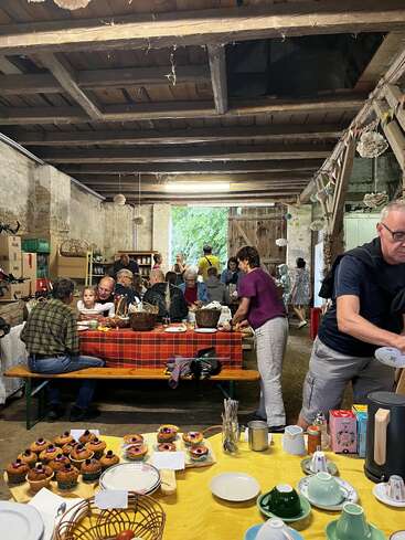 Ein gemütliches Beisammensein in einer rustikalen Scheune mit Menschen, die Essen und Gespräche genießen. Cupcakes, Teller und Tassen stehen auf den Tischen und schaffen eine warme, gemeinschaftliche Atmosphäre.