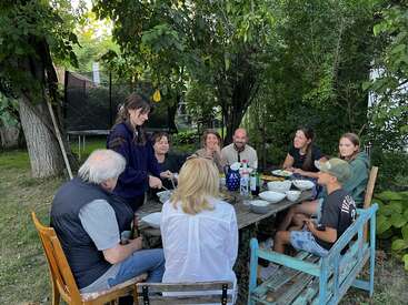 Eine Gruppe von Menschen genießt ein gemeinsames Essen im Freien an einem rustikalen Holztisch, umgeben von Bäumen und Grünzeug. Alle scheinen glücklich und entspannt zu sein.