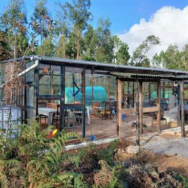Eine rustikale Gewächshauskonstruktion mit Glasfenstern und Holzrahmen. Drinnen stehen Tische und Stühle, umgeben von Bäumen, Pflanzen und einem blauen Himmel. Eine friedliche Umgebung.