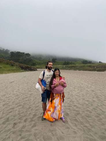 Um casal está junto em uma praia de areia com colinas enevoadas e árvores ao fundo, vestido casualmente, carregando bolsas e segurando uma criança pequena.