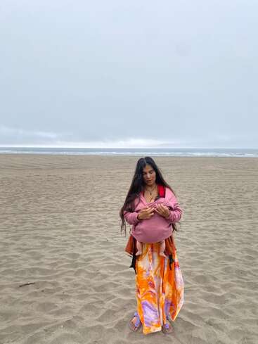 Uma mulher está em uma praia de areia sob um céu nublado, vestindo uma saia colorida e um suéter rosa, segurando um bebê em um carrinho.