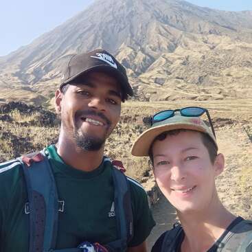Two people smiling and posing for a selfie outdoors, wearing casual hiking attire and hats, with a large, rocky mountain dominating the background under clear skies.