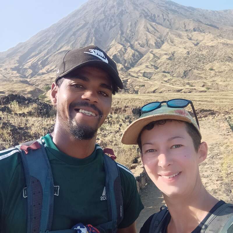 Duas pessoas sorrindo e tirando uma selfie ao ar livre, vestindo roupas casuais de caminhada e chapéus, com uma grande montanha rochosa dominando o fundo sob um céu claro.