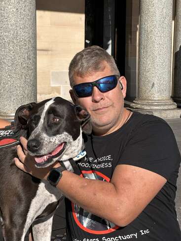 A man wearing sunglasses and a black t-shirt hugs a happy black-and-white dog outside a building with stone columns, both enjoying the sunshine together.