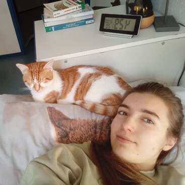 A young woman reclines on a bed, smiling slightly, while a ginger and white cat rests behind her head. Books and a digital clock are nearby.