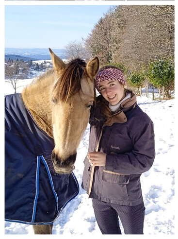 A smiling person stands closely beside a light brown horse in a snowy landscape, both warmly dressed. Trees and distant hills complete the peaceful winter scene.