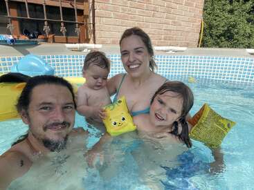 Una feliz familia de cuatro miembros disfruta de un día soleado en una pequeña piscina en el patio trasero. Los padres sonríen mientras sus hijos pequeños juegan con flotadores y juguetes.