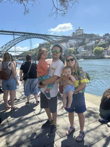 A family of four poses by the riverside with a scenic bridge and cityscape in the background, enjoying a sunny day among other tourists.