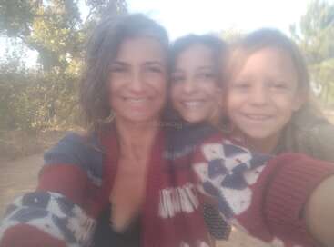 Une femme et deux enfants sourient ensemble dans un cadre extérieur, vêtus de pulls confortables. La photo est douce et floue, créant une atmosphère chaleureuse, joyeuse et détendue.