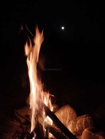 Un feu de camp crépitant brûle vivement, entouré de pierres, ses flammes dansant dans le ciel nocturne. La lune brille doucement au-dessus, illuminant l'obscurité paisiblement.