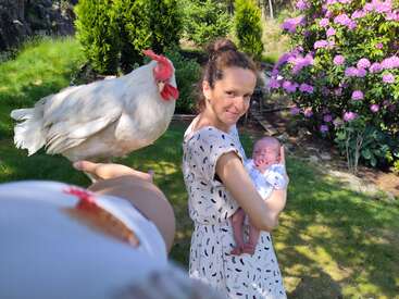 Eine Frau hält ein neugeborenes Baby in einem sonnigen Garten und steht neben jemandem, der ein weißes Huhn hält. Üppiges Grün und blühende lila Blumen umgeben sie.