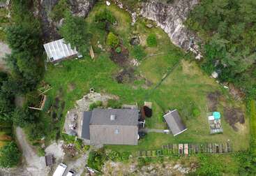 Dieses Luftbild zeigt ein Haus mit Garten, Gewächshaus, Schuppen, mehrere Gartenbeete, Rasen, Bäume, felsiges Gelände, Wege, ein Fahrzeug und die natürliche Umgebung.