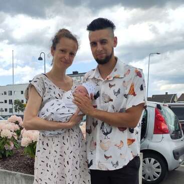 Ein glückliches Paar steht draußen, lächelt und hält sein neugeborenes Baby im Arm. Beide Eltern tragen gemusterte Kleidung. Es ist ein fröhlicher Familienmoment an einem bewölkten Tag.