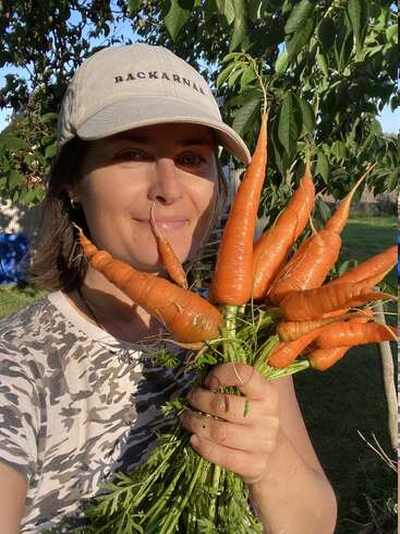 A smiling woman in a cap holds a fresh bunch of homegrown carrots with dirt still on them, standing outside in a sunlit, green garden.