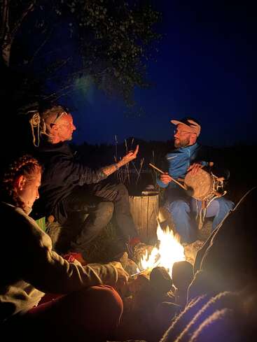 Four people sit around a campfire at night. One person plays a drum, another gestures while talking, creating a warm, intimate, and peaceful outdoor scene.
