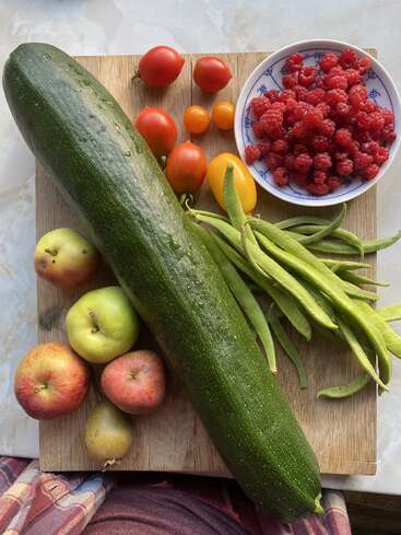 A vibrant assortment of freshly harvested produce: large zucchini, tomatoes, yellow beans, apples, a kiwi, and a bowl of bright red raspberries on a wooden board.