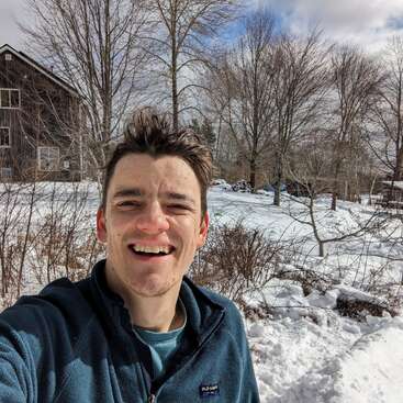Ein Mann lächelt für ein Selfie draußen in einer verschneiten Landschaft. Vor dem hellen Winterhintergrund sind kahle Bäume und ein rustikales Holzhaus zu sehen.