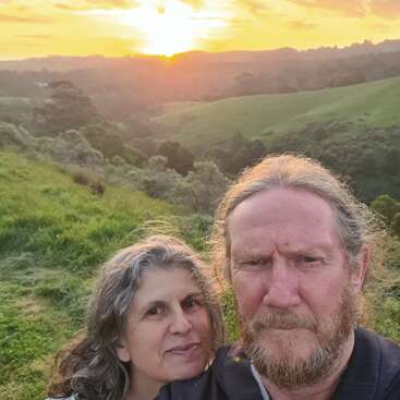A couple stands together on a grassy hillside, capturing a selfie as the sun sets over green rolling hills and forests, creating a peaceful atmosphere.