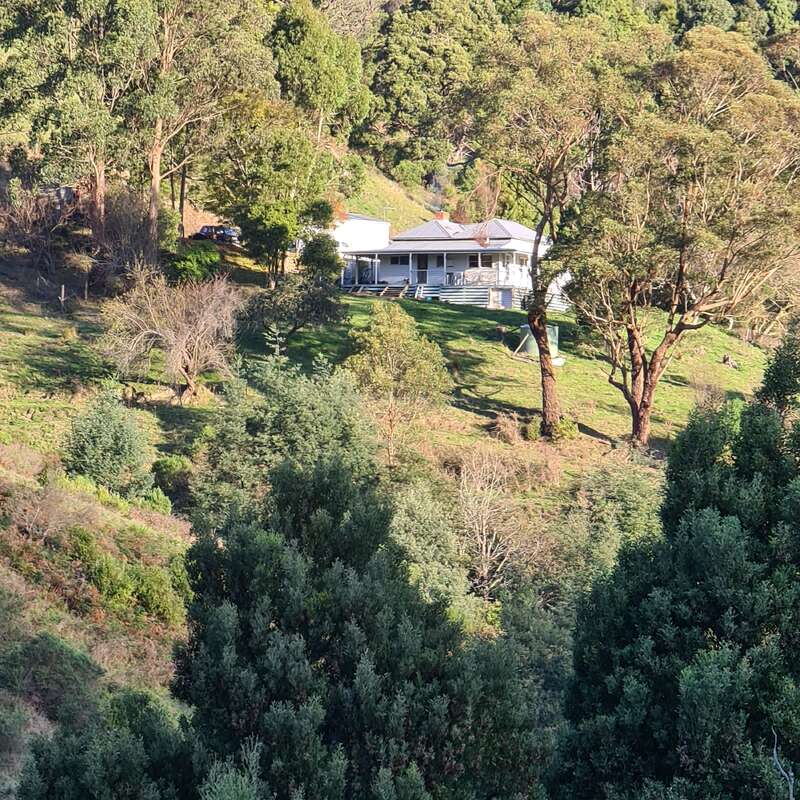 The image depicts a serene white house with a wraparound porch, nestled among trees on a hillside, surrounded by lush greenery and a vibrant natural landscape.