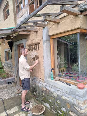 Ein Mann verziert eine irdene Wand mit dem Wort "माया" (was auf Hindi "Liebe" bedeutet) vor einem rustikalen Haus mit natürlichen Materialien und einfachen Werkzeugen.