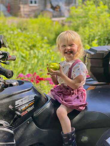 Ein lächelndes junges Mädchen mit blonden Haaren sitzt auf einem schwarzen Quad und hält einen grünen Apfel in der Hand. Sie trägt draußen einen rosa Rock, ein weißes Hemd und Gummistiefel.