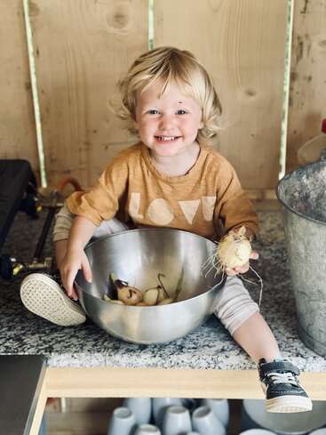 Ein lächelndes Kleinkind mit blonden Haaren sitzt auf einer Theke und hält eine Zwiebel in der Hand. Es trägt ein T-Shirt mit der Aufschrift „LOVE“ und sitzt neben einer Metallschüssel voller Zwiebeln.
