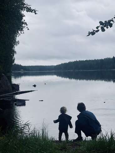Ein Kind und ein Erwachsener stehen an einem ruhigen See unter bewölktem Himmel, umgeben von Bäumen, und genießen gemeinsam einen stillen, friedlichen Moment in der Natur.
