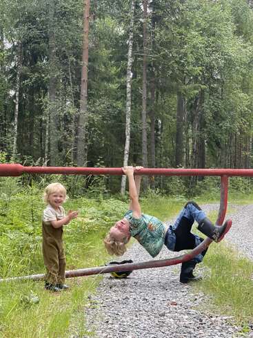 Zwei Kinder spielen draußen an einem roten Metalltor; eines hängt kopfüber, während das andere lächelnd daneben steht, inmitten einer üppig grünen Waldlandschaft.