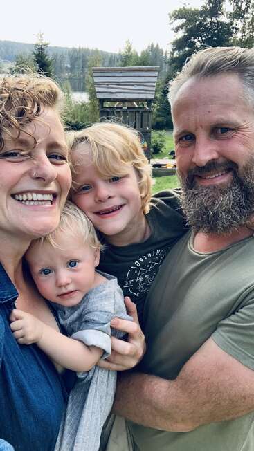 Eine lächelnde vierköpfige Familie posiert eng beieinander im Freien. Zwei Erwachsene und zwei kleine Kinder genießen einen sonnigen Tag, hinter ihnen sind Grünflächen und ein Spielgerüst aus Holz zu sehen.