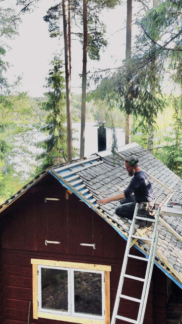 Un hombre está sentado en un tejado empinado, trabajando con herramientas para reparar o instalar tejas, rodeado de árboles altos y un pintoresco paisaje natural.