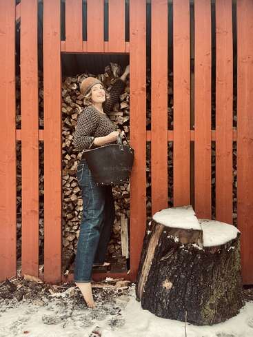 Una mujer sonriente, con sombrero y jersey, recoge leña de una pila apilada. Está descalza sobre la nieve, junto a un gran tocón de árbol.