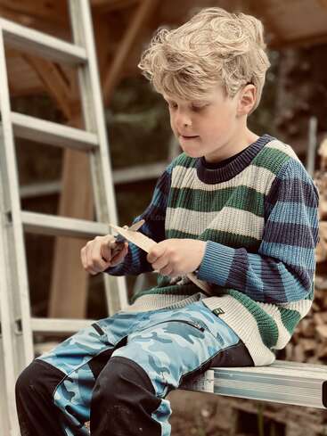 Un niño rubio está sentado en una escalera metálica al aire libre, vestido con un jersey a rayas y unos pantalones de camuflaje azules, tallando madera con gran concentración.