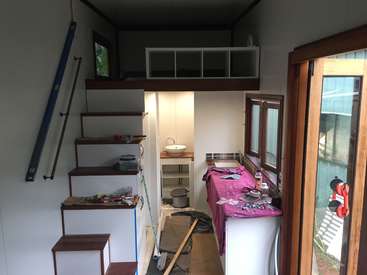 Das Bild zeigt das Innere eines kleinen, unfertigen Raums mit weißen Wänden und hölzernen Akzenten, mit einer Treppe, einem Waschbecken und einer Werkbank, auf der Werkzeuge und Materialien verstreut sind.