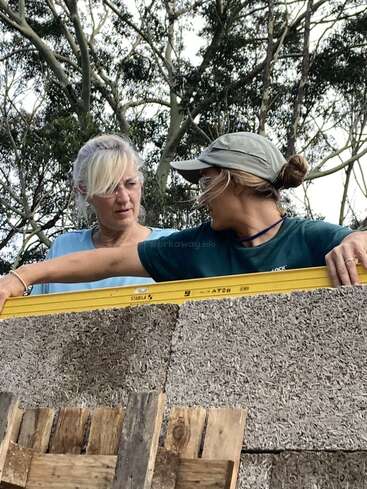 Two women are working together outdoors, using a yellow level tool to check the alignment of a wall made from natural building materials, surrounded by trees.
