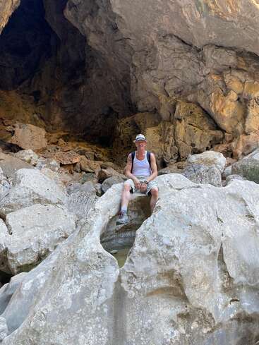 Das Bild zeigt einen Mann, der auf einem großen Felsen vor einer Höhle sitzt. Er trägt ein weißes Tanktop, Shorts und einen Hut und hat einen Rucksack auf dem Rücken.