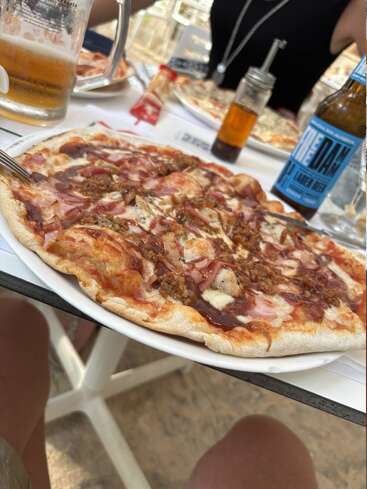 Das Bild zeigt einen Tisch, auf dem eine Pizza, Bier und andere Lebensmittel liegen, wahrscheinlich in einem Restaurant oder Café im Freien.