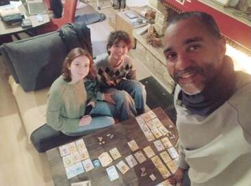Three people enjoying a cozy game night, smiling together beside a fireplace. They’re playing a tabletop card game, surrounded by a relaxed and warm home atmosphere.
