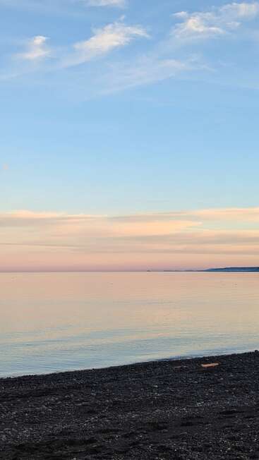 Eine ruhige Strandszene bei Sonnenuntergang. Sanfte Pastellfarben färben den Himmel und spiegeln sich auf dem ruhigen Meer wider, mit einer Kieselstrandküste im Vordergrund. Friedliche Atmosphäre.