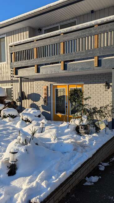 Ein Haus mit gelben Doppeltüren, einem verschneiten Vorgarten, Winterpflanzen, einem Balkon mit Holzgeländer und strahlendem Sonnenschein, der die Winterszene beleuchtet.
